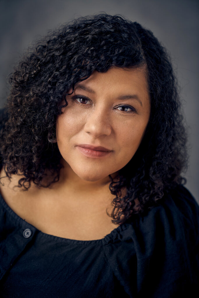 A photo of Zilla Jones. She is an Black woman with shoulder-length curly hair and brown eyes. She is wearing a navy-blue top and looking into the camera with a slight smile. 