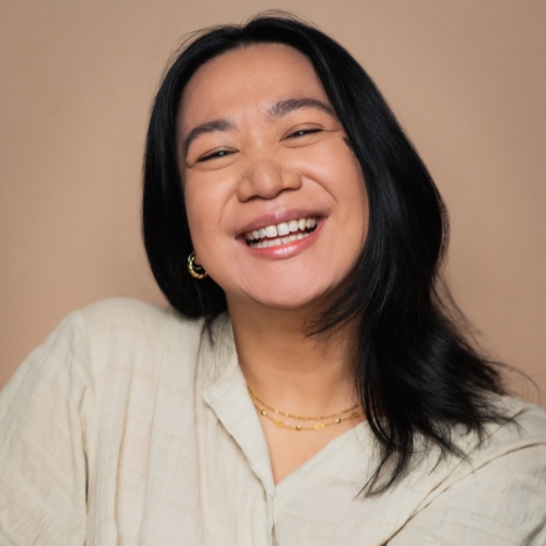 A photo of Kamila Sediego. She is a woman of Filipinx descent with shoulder-length black hair. She is standing in front of a neutral-coloured wall and smiling into the camera. 