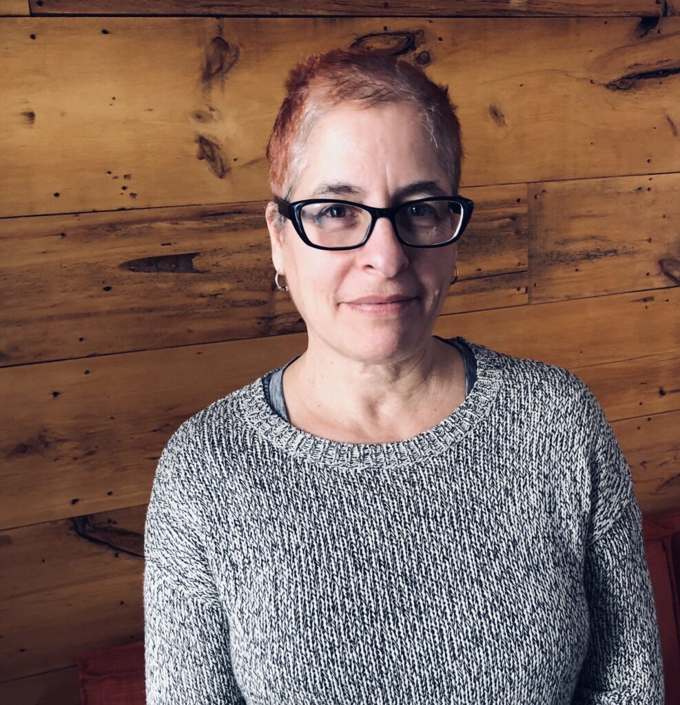 A photo of Leila Marshy. She is a light-skin-toned woman wearing glasses and a grey sweater, standing against a wooden background.