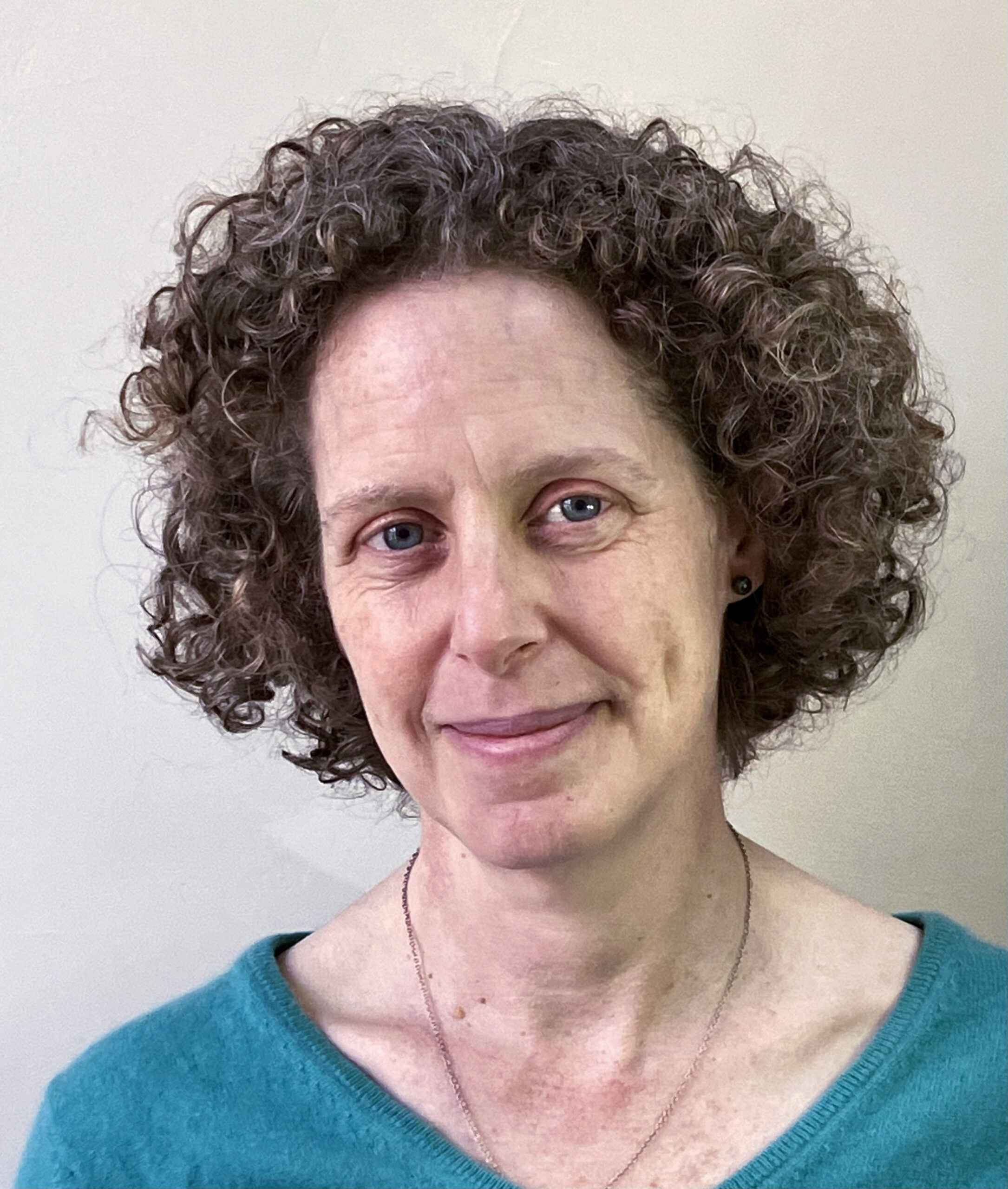 A photo of Megan J. Elias. A light-skin-toned person with short curly hair wearing a teal sweater and a delicate necklace, smiling softly against a neutral background.