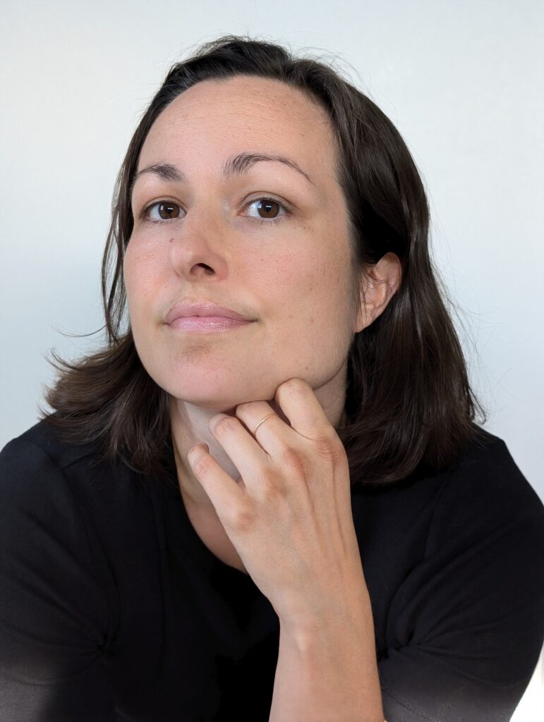 A photo of Nadia Staikos, a light-skin-toned woman with dark hair. She is resting her chin on her hand and looking into the camera. 
