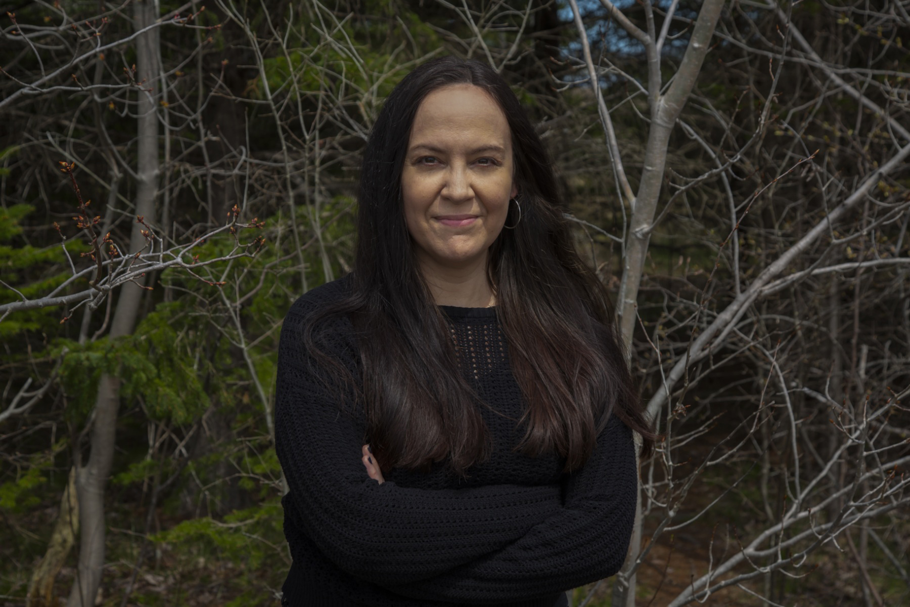 A photo of author Vanessa F. Penney. She is a light-skin-toned woman with long dark brown hair. She is wearing a black sweater and standing in front of a wooded area smiling into the camera with her arms crossed.