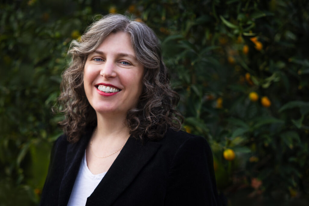 A photo of Rebecca Morris, a light-skin-toned woman with curly salt-and-pepper hair wearing a black blazer and white top and smiling into the camera.