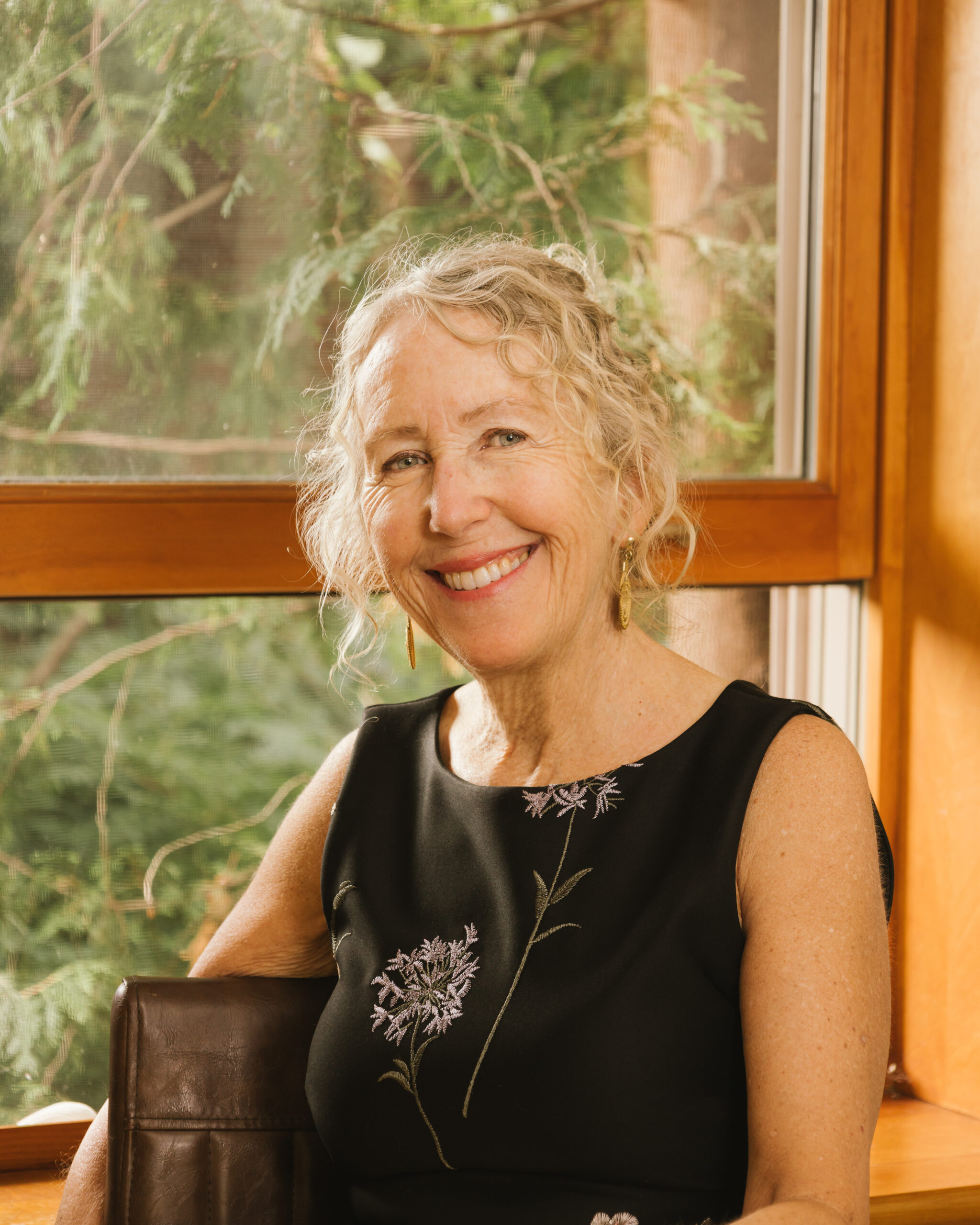 A photo of Susan Grundy. She is a light-skin-toned woman with curly blonde hair. She sits indoors beside a window with sunlight streaming in. She wears a sleeveless black dress with embroidered purple flowers and gold earrings, with green foliage visible outside the window.