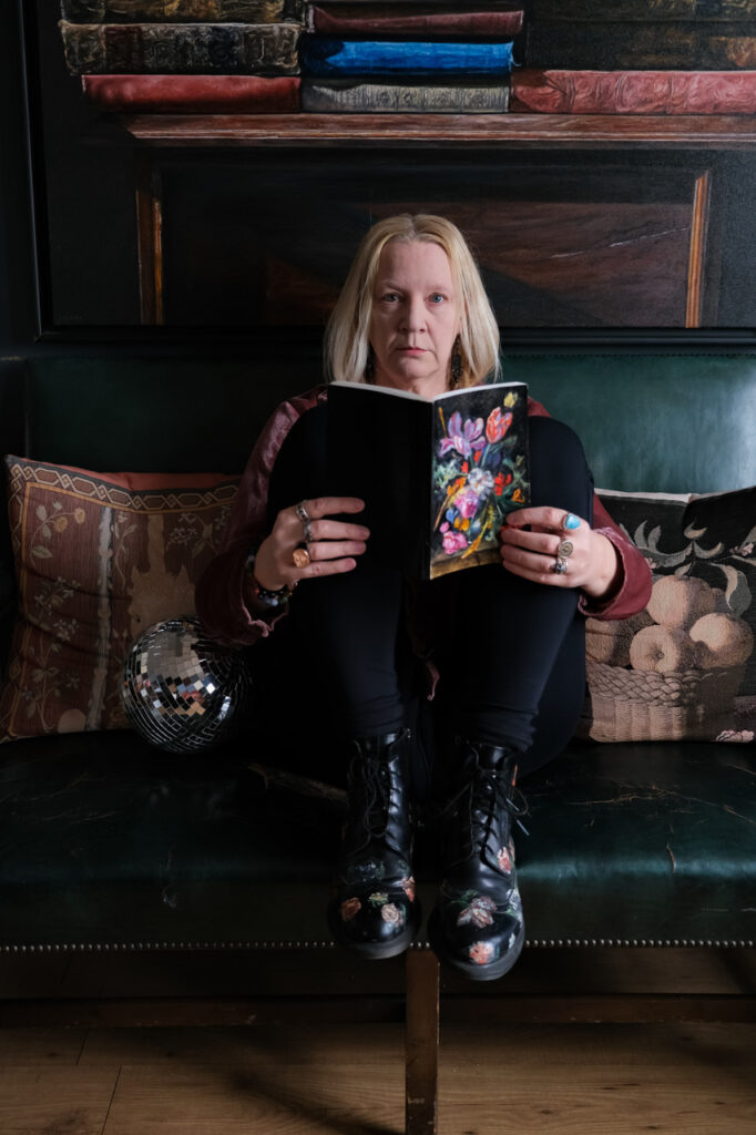 A photo of Shawna Lemay. She is a light-skin-toned woman with blonde hair. She is sitting with her knees tucked up to her chest on a green velvet couch with a disco ball and decorative pillows behind her, holding a flower-patterned notebook open. She is looking into the camera.