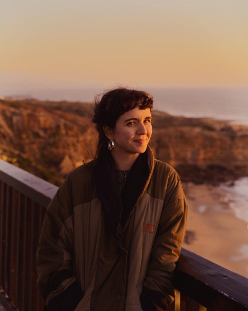 A colour photo of Joana Mosi standing by a wooden railing overlooking a rocky beach at sunset, wearing a dark jacket and smiling slightly.