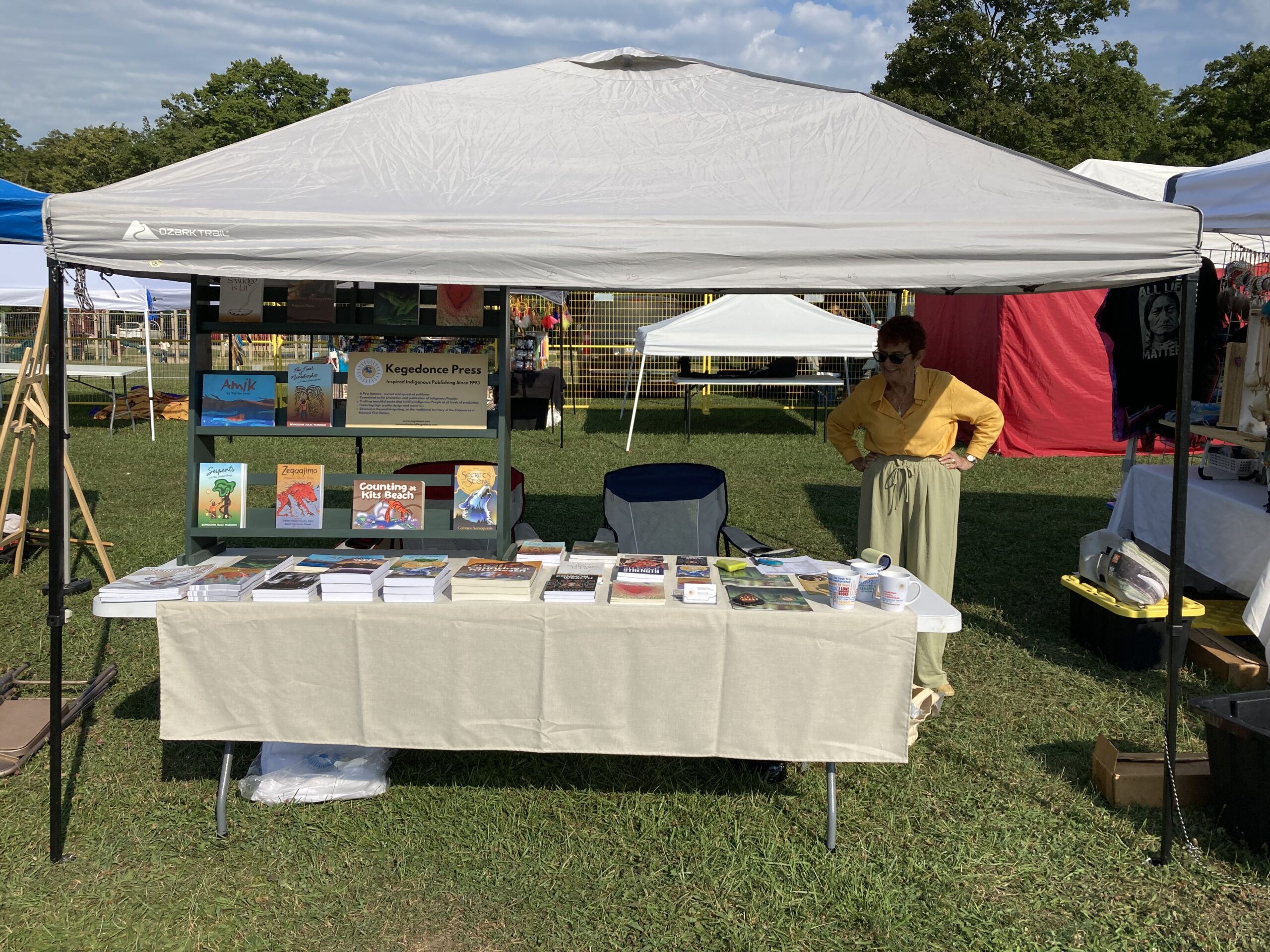 A photo of Kegedonce Press's administrator Christy Telford at Neyaashiinigmiing Pow Wow. The photo shows Christy standing beside a table of books