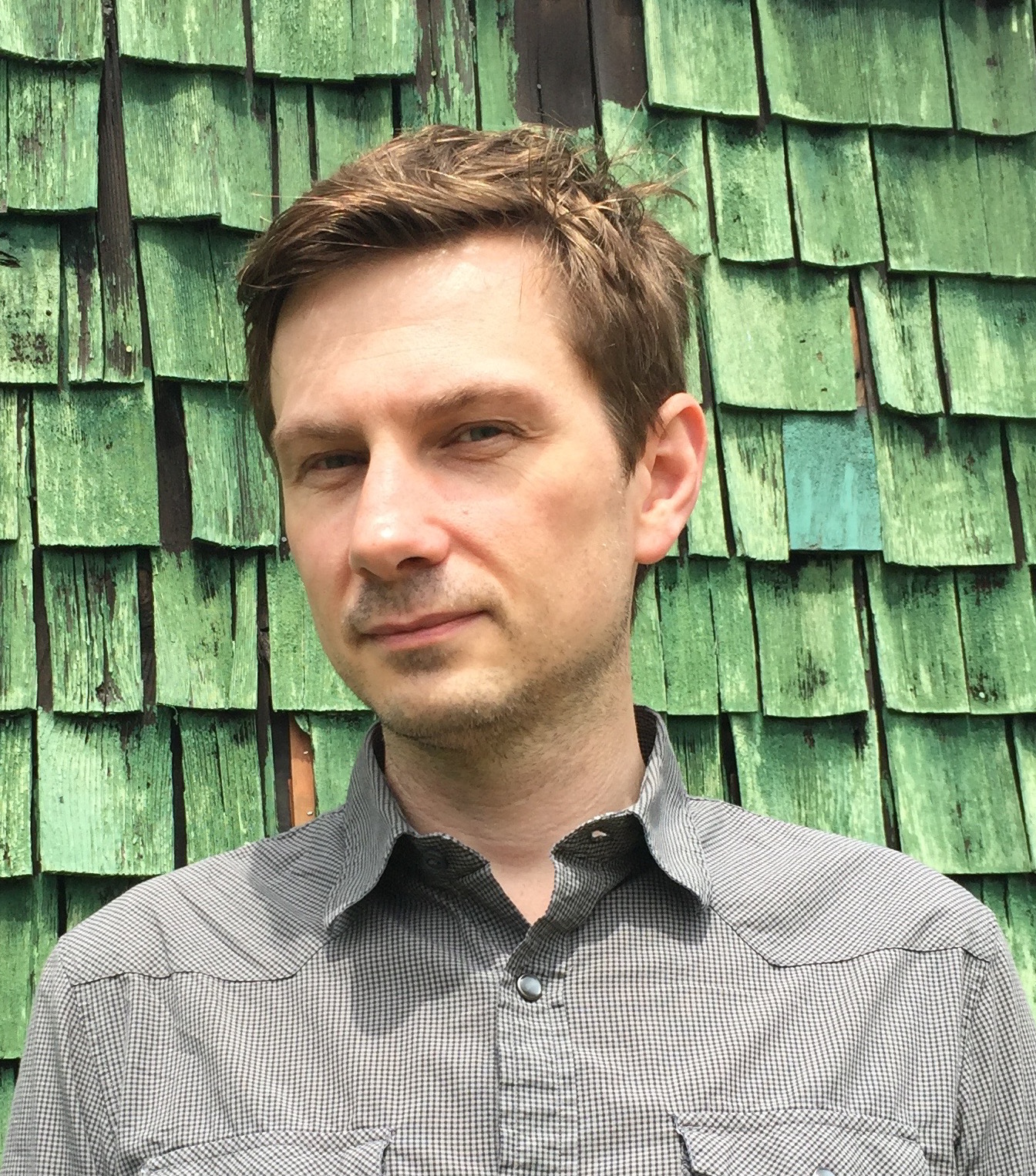 A photo of Greg Rhyno. He is a light-skin-toned man with short sandy-blonde hair. He is standing in front of  a wall of green shingles and looking into the camera. 