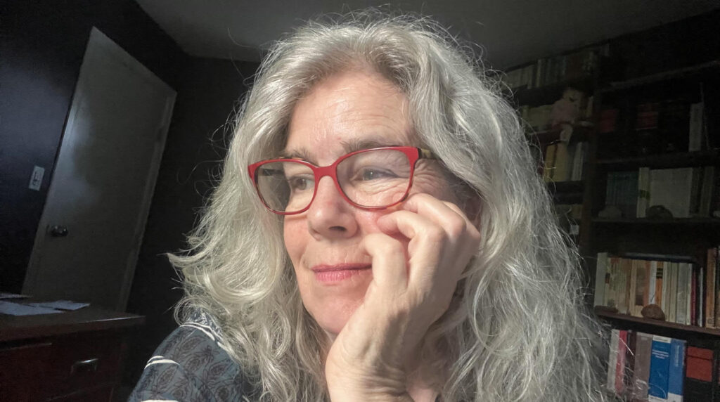 A photo of Alison Acheson. She is a light-skin-toned woman with long grey hair and large red-framed glasses. She is propping her head up with one hand and looking to the side with a slight smile. 