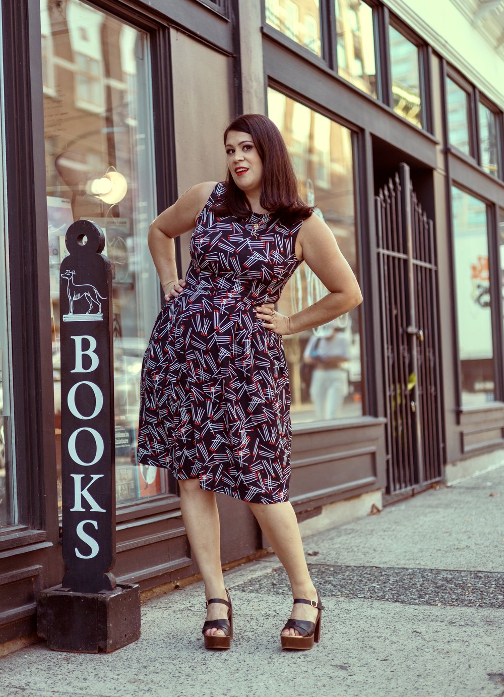 A photo of Dina Del Bucchia. She is a light skin-toned woman with shoulder-length brown hair. She is wearing a patterned knee-length dress and open-toed heels and standing on the sidewalk beside a sign that reads "books."